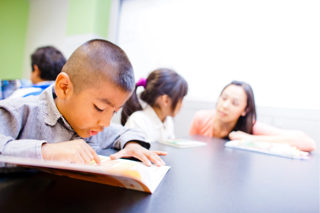 Young boy reading a book at a table. Another student and teacher nearby