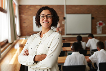 A female teacher poses with her arms crossed in her classroom with students behind her